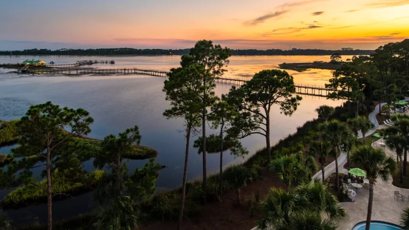 Aerial view of water, trees, bridge, buildings, pool, and chairs, with a colorful sunset sky.