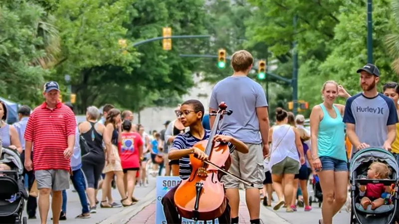 A crowd of people are walking on a sidewalk and one of them is playing a cello.