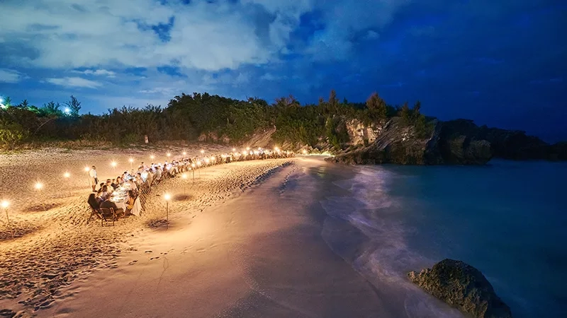 A long table with white cloth and chairs at night on the beach with lit candles on sand.