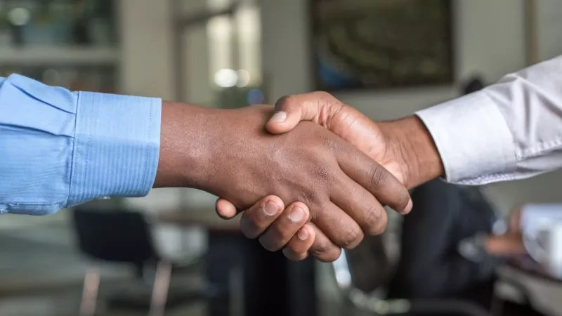 Two people shake hands in an office while another person sits nearby and a cup rests on a table.