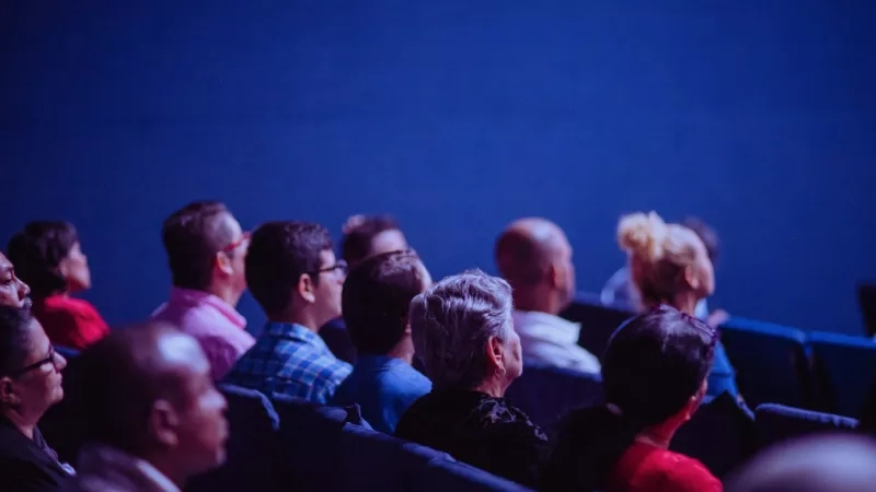 Audience in an auditorium with blue walls and chairs, watching a presentation.