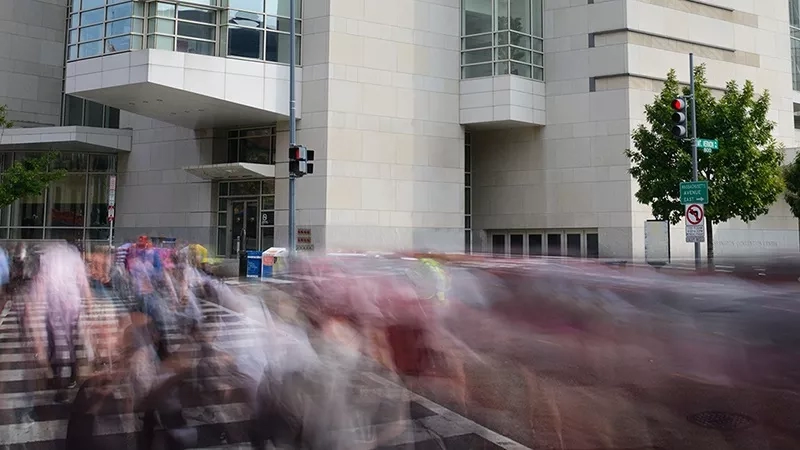 People walk across the street in front of a large building with glass windows and a tree in the corner.
