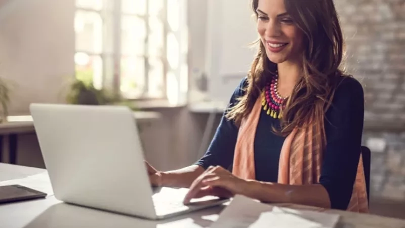 A woman is sitting on a chair and working on a laptop at the desk.