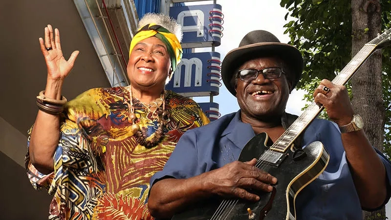 Two musicians in front of the Blues Museum in Clarksdale, Mississippi, waving and holding a guitar.