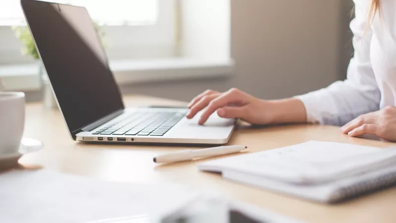 A person working on a laptop at a desk with papers and a pen.