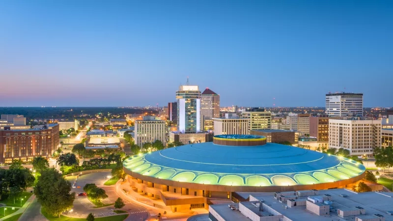 Aerial view of downtown Tulsa at dusk with illuminated skyscrapers, lush landscaping, and a serene ambiance.