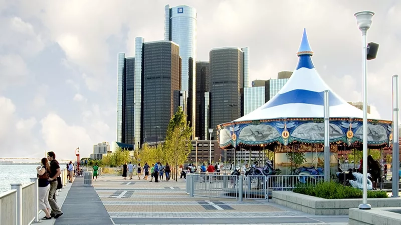 Detroit International Riverfront has a carousel with people walking near tall buildings in the background.
