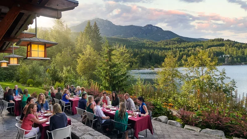 Group of people dining on an outdoor patio with a lake and mountains in the background.