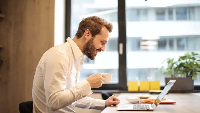 Man working on laptop and holding coffee cup in virtual networking event at office.