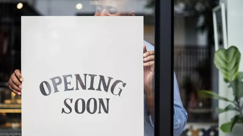 Man putting up an "Opening Soon" sign on a glass door in front of a blurred store interior.