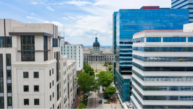 A cityscape of Columbia with the South Carolina State House and Synovus building, surrounded by greenery.