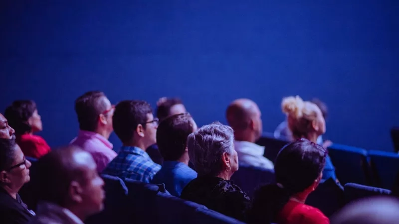 A group of people sit in a dimly lit theater, watching a show or movie.