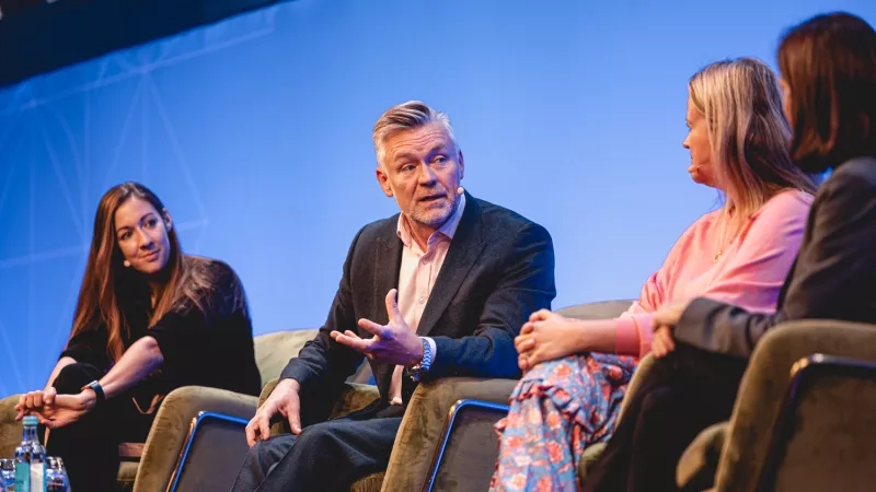 Four people sit on chairs in a conference room, with a man gesturing and a woman listening attentively.