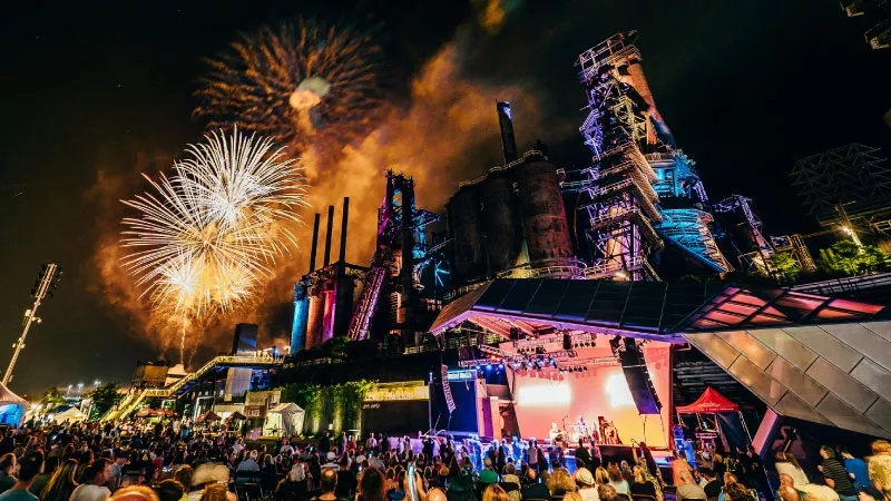 A crowd gathers as fireworks light up the sky above the Pittsburgh skyline at night.