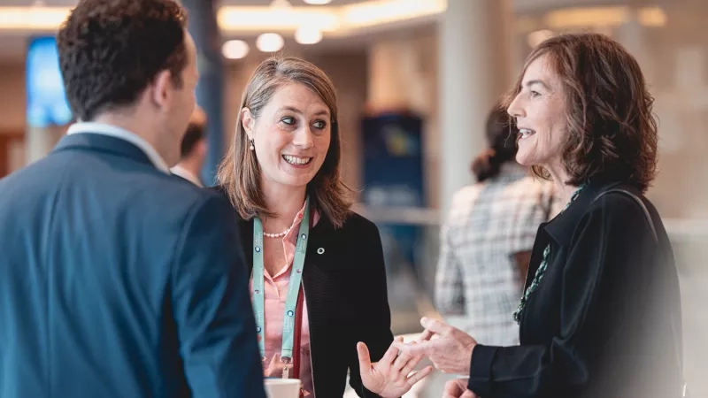 Two women and a man converse at a round table at a conference with others in the background.