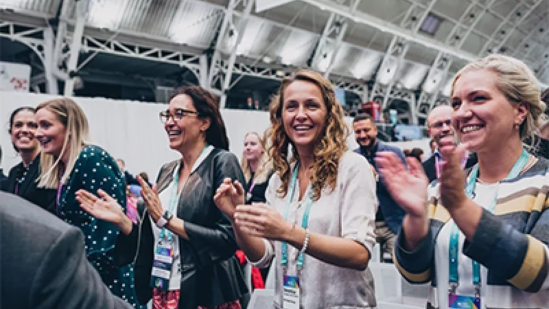Several women wearing lanyards are clapping their hands in an indoor event space with steel architecture.