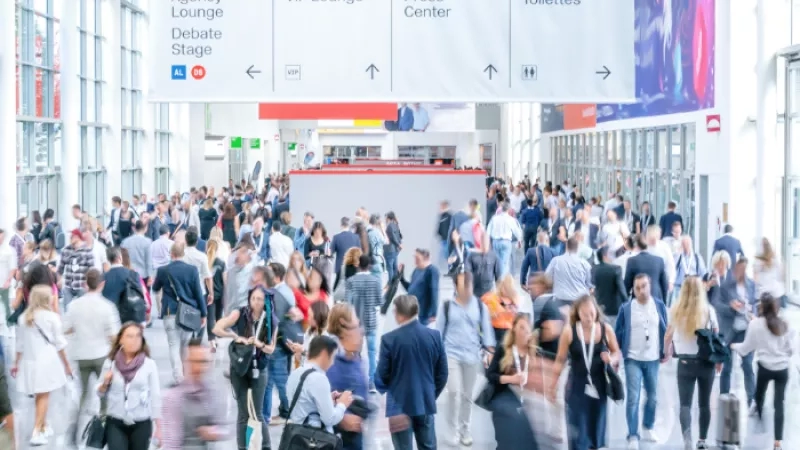 A large crowd of people at a convention with a large white sign hanging from the ceiling.