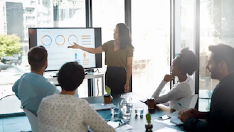 Woman giving a presentation in a conference room with others watching her in front of a TV.