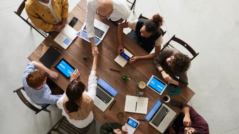 Group of people gathered around a wooden table with laptops, tablets, and notebooks.