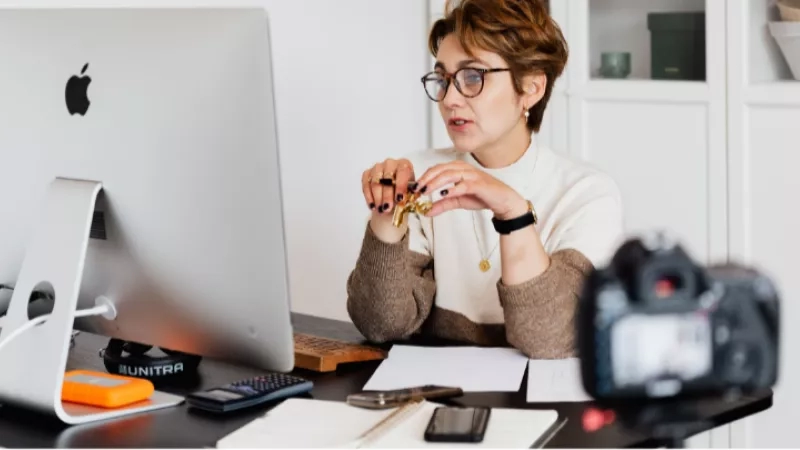 A woman with curly hair is sitting on a desk in front of an Apple computer.