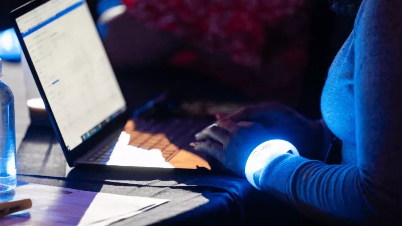 A woman wearing a wristband with a light works on a laptop at a table in the dark.
