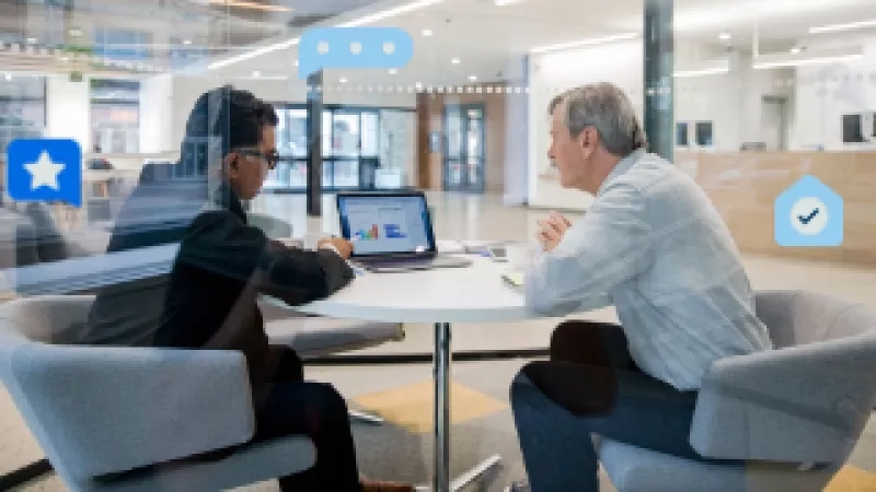 Two people are sitting on chairs facing each other and discussing something while a laptop is on the table.