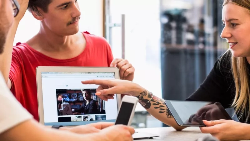 Three people in a meeting room with a laptop, tablet, phone, and headphones on the table.