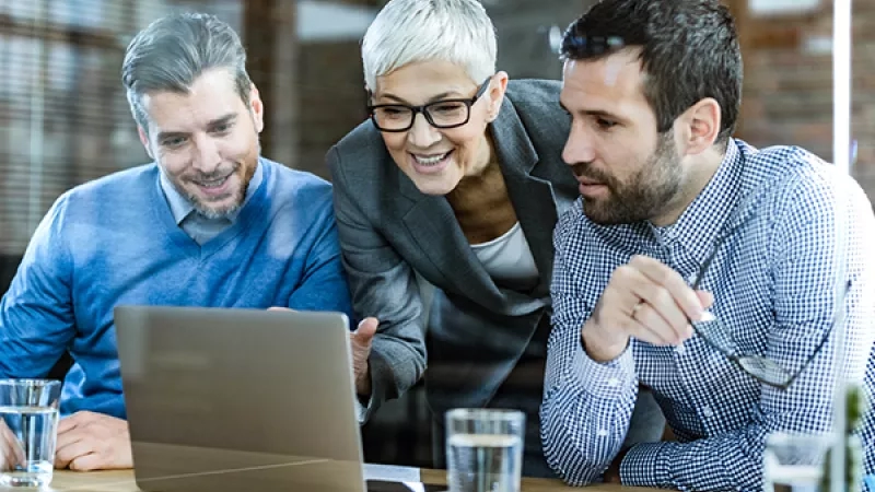 Three business people looking at a laptop on a table with glasses of water in front of them.