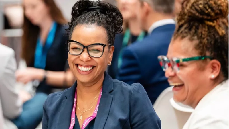Two women laughing at a conference with others in the background.