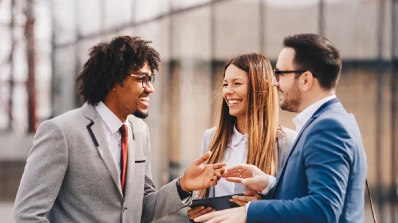 Three professionals in formal attire stand in a line, smiling and talking to each other outside.