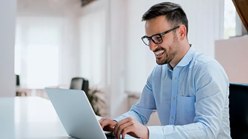 A smiling man wearing glasses works on a laptop on a desk in a modern office.