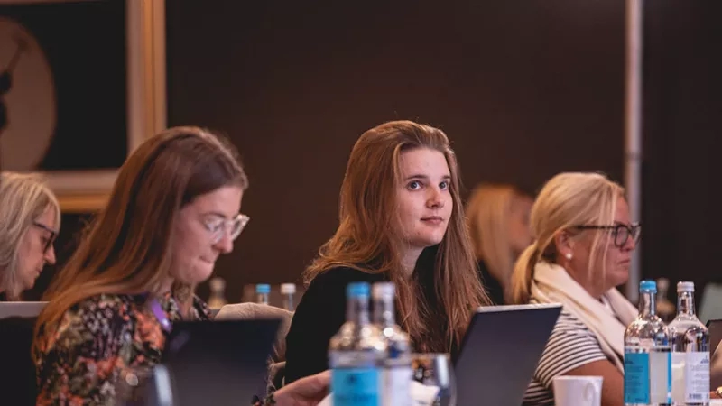 Group of women at a conference table with laptops, bottles, and glasses of water in front of them.