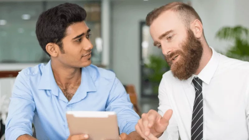 Two men in an office setting, one holding a tablet, engaging in a conversation.