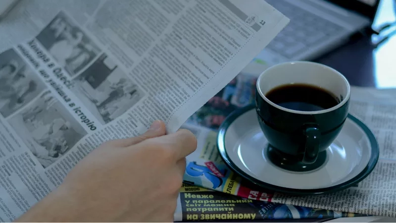 A hand holding a newspaper with a coffee cup and saucer on a table.