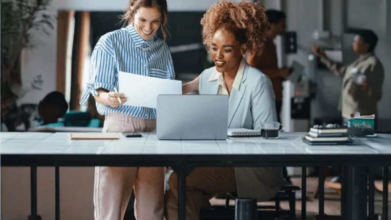 Two women are laughing and reviewing documents with a laptop in front of them in a casual office environment.