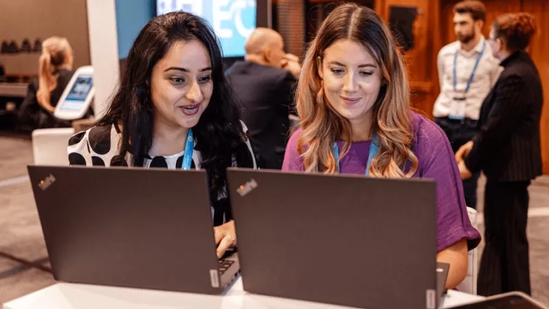 Two women with laptops at a conference with a large screen and other people in the background.