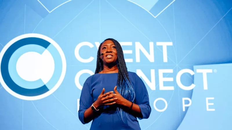 A woman is giving a speech on stage in front of a blue backdrop with text and logos.