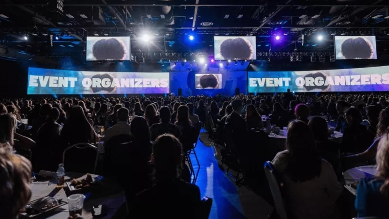 Many people sitting in a conference hall with a big screen and text "Event Organizers" on it.