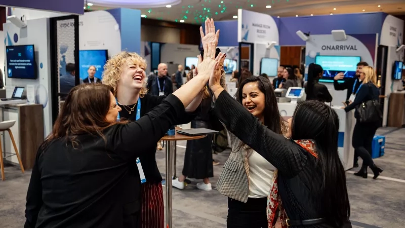 Four women high-fiving each other at a conference with a sign that reads "Onarrival" in the background.