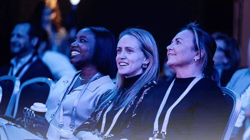 Three women laughing at an event, with one holding a coffee cup and others in the background.