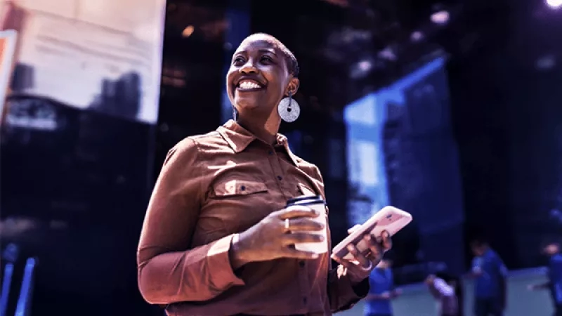 A person in a brown shirt holds a coffee cup and a smartphone, standing in a modern indoor space