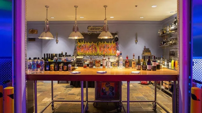 A bar counter with multiple bottles on display and a painting of fire on the wall.