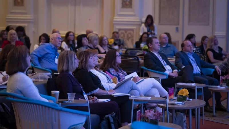 A large group of people sitting in chairs in a conference room listening to a speaker.