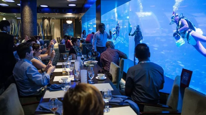 People are dining at a table in front of an aquarium with divers swimming in the background.