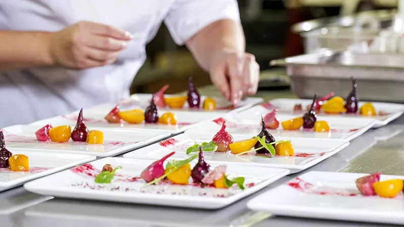 A chef is plating a dish with various vegetables on a white plate in a kitchen.