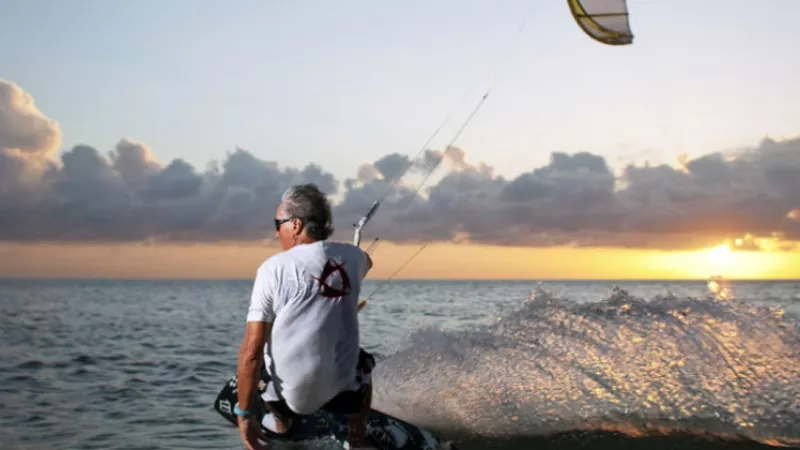 A man is kitesurfing on the ocean as the sun sets in the background.