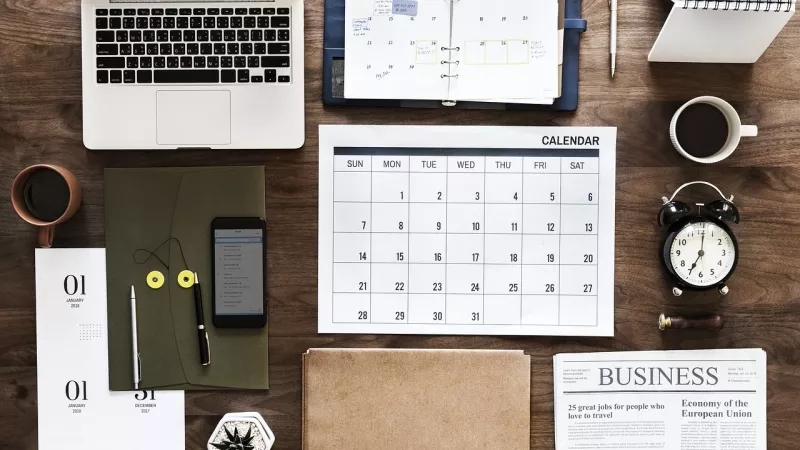 Laptop, calendar, alarm clock, business newspaper, and coffee on a wooden table.