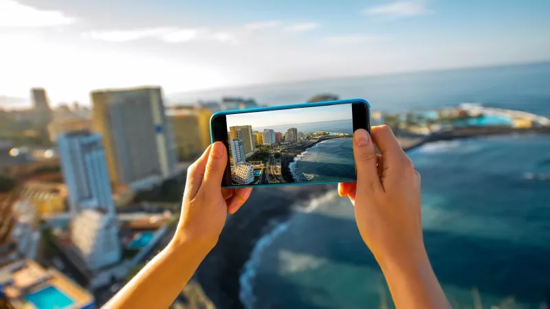 Person holding smartphone over beach with a view of a city in the background, capturing the scenery.