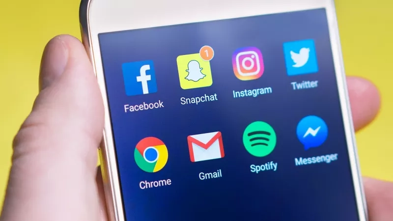 A person holds a phone displaying app icons like Facebook, Snapchat, and Spotify on a yellow background.