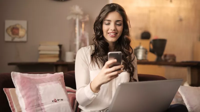 A woman is smiling while holding her phone and using her laptop on the couch.